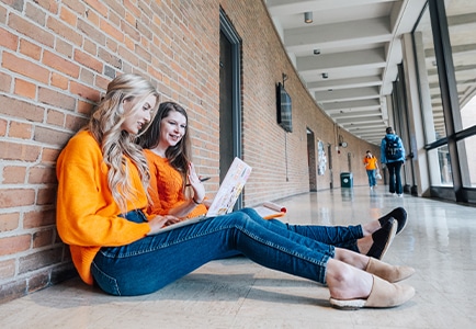 Two female students wearing orange sit in the hallway of the College of Communications