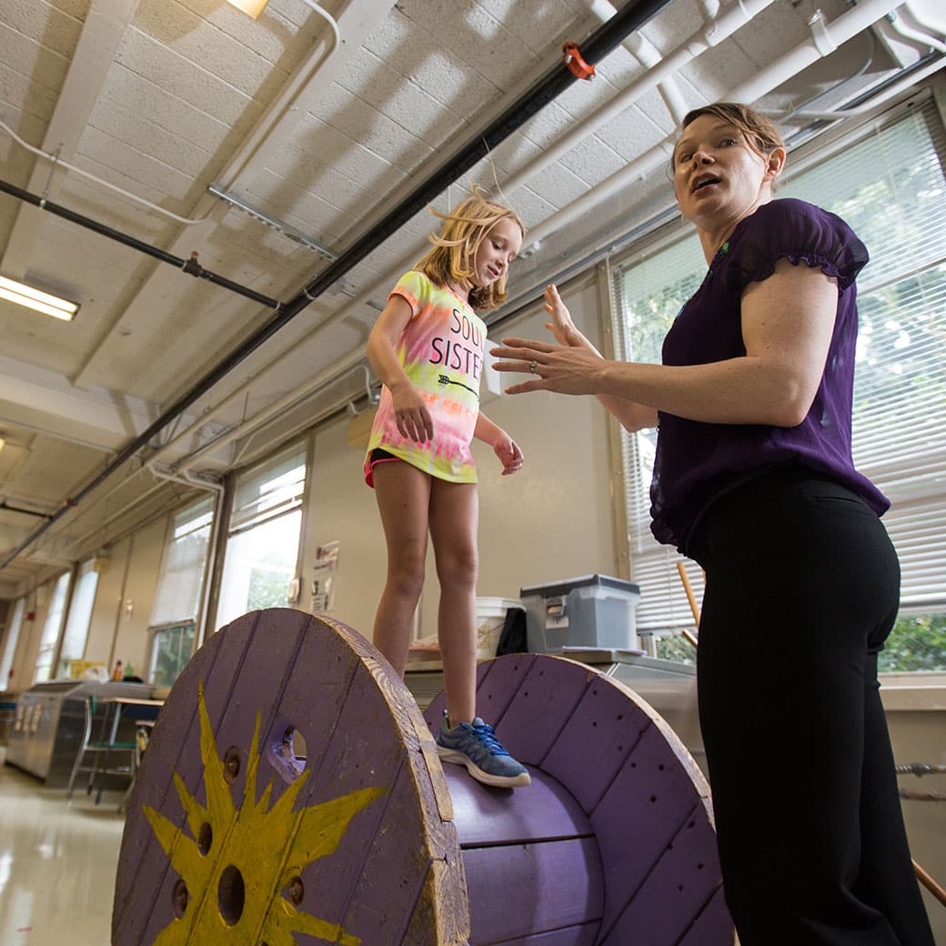 A woman works with a young girl who is standing on a purple wheel