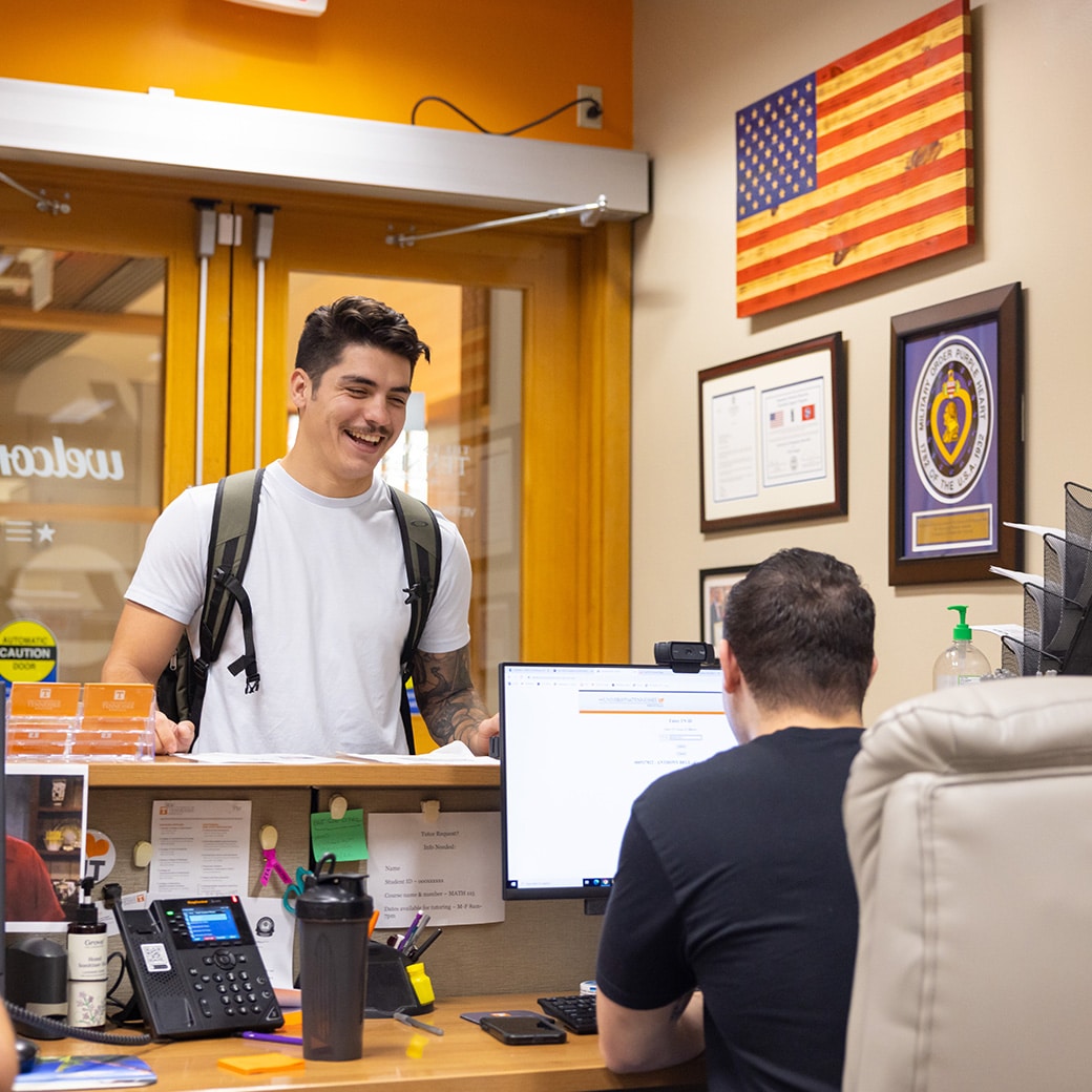 A male stands at a welcome desk and chats with a person seated at a desk in the Veterans Success Center