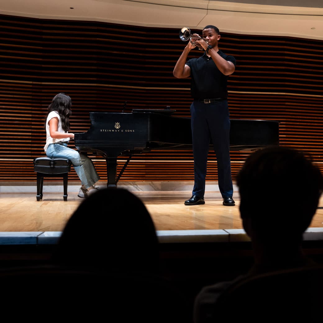 A student plays the piano in the background as another student plays the trombone in the foreground