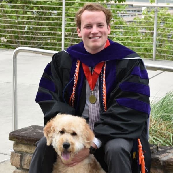 Zach Campbell in commencement regalia posing with a blonde dog