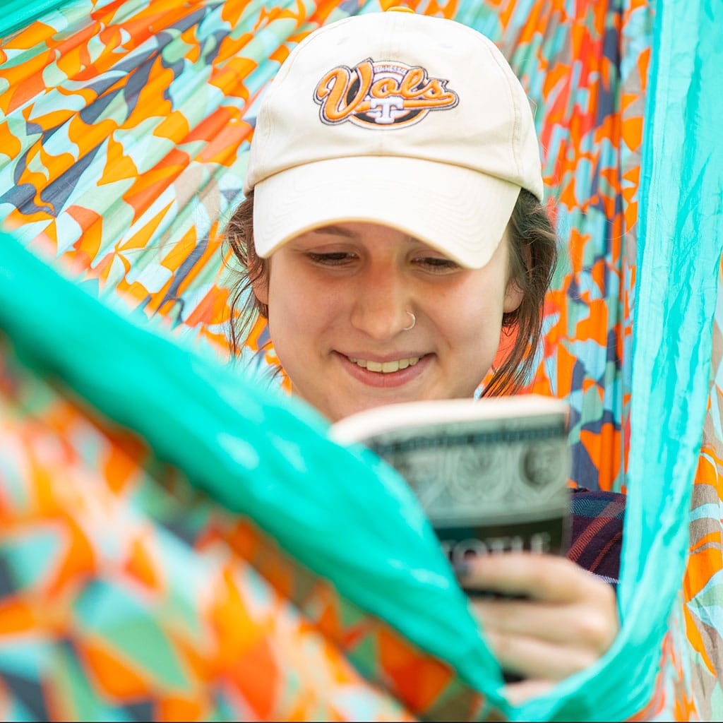 A student wearing a baseball cap relaxes in a bright orange and teal hammock, reading a book