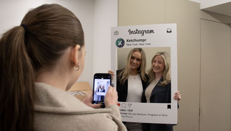 Two blonde females pose with a frame that looks like an Instagram post as a brunette female takes a photo on her cell phone