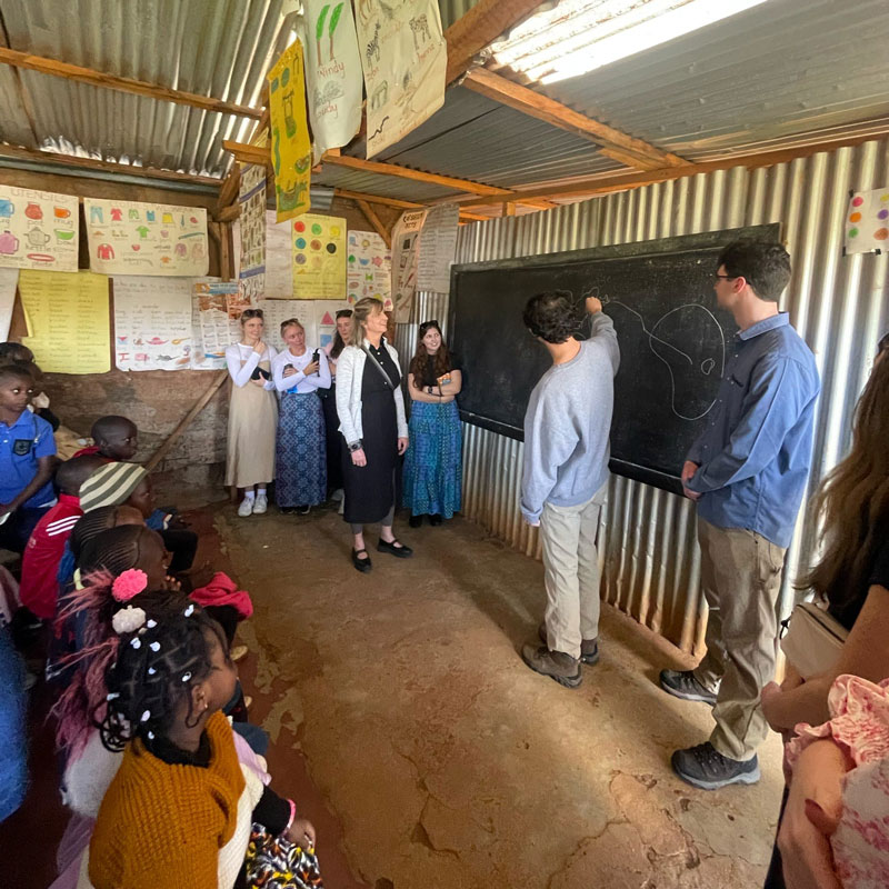 greenberg-oster-in-kenya-800x800-2 Jiro Greenberg-Oster points to a chalkboard with a drawing while schoolchildren look on.