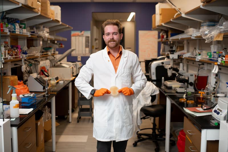 myers-tyler-lab-800x533-2 Tyler Myers stands in the middle of the Goodrich-Blair Lab, wearing a white lab coat, and holding a petri dish with an orange Power T.