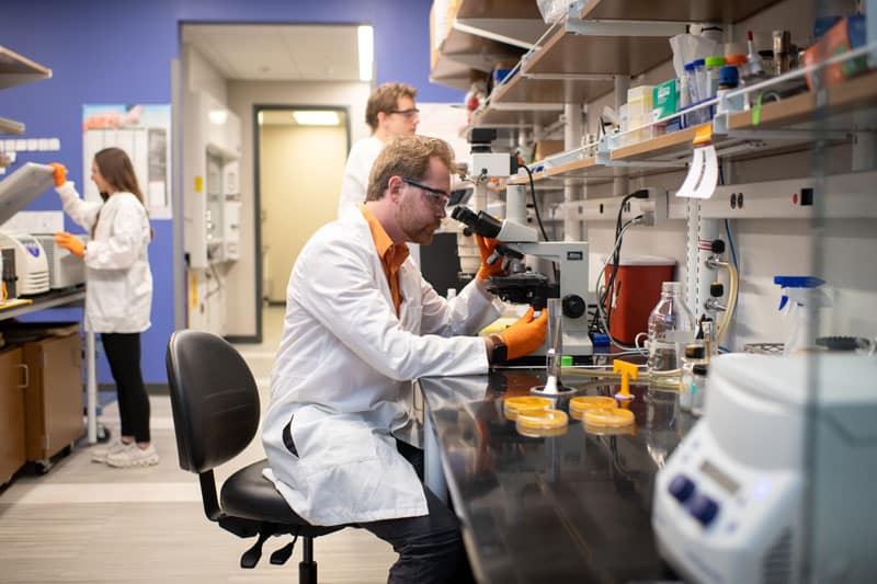 myers-tyler-lab-microscope-800x533 Tyler Myers looks through a microscope. In the background, two other students are working in the lab.