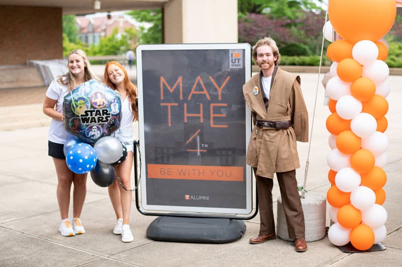 myers-tyler-saa-may-the-4th-800x533 Tyler Myers, dressed as a Star Wars Jedi, stands to the right of a sign that says May the 4th Be with You. Two more students stand to the left of the sign, holding Star Wars-themed balloons.
