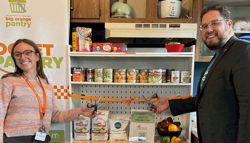 Blake Weiss and a woman stand in front of a pantry stocked with food with an orange ribbon across it. Blake holds scissors, about to cut the ribbon.