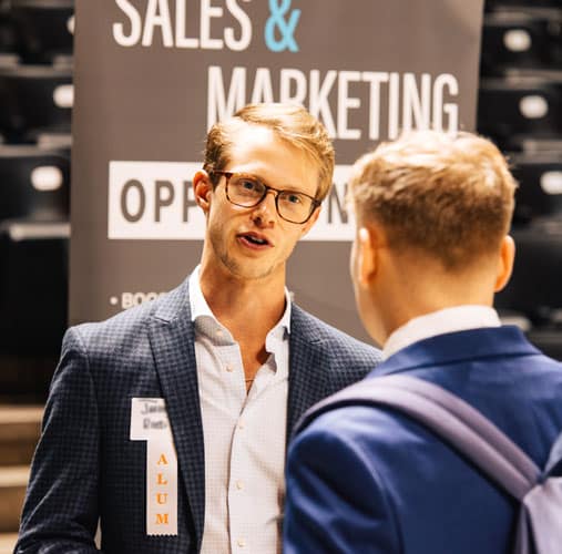 fye-2025-gallery-career-fair Two men talking during a career fair. In the background is a banner that says Sales and Marketing Opportunities.