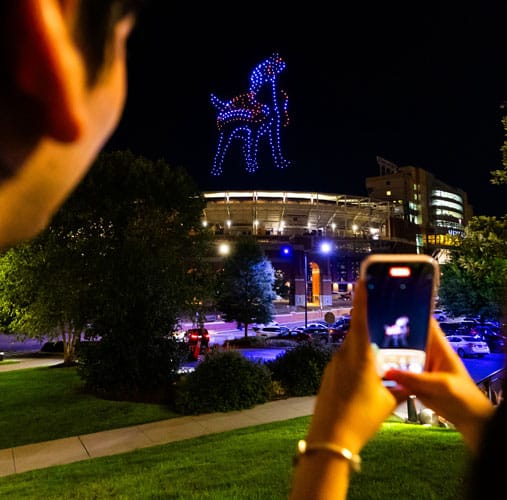 fye-2025-gallery-drone-over-neyland Two hands holding a cell phone, taking a photo of Neyland Stadium with a Smokey light projection above it at night.