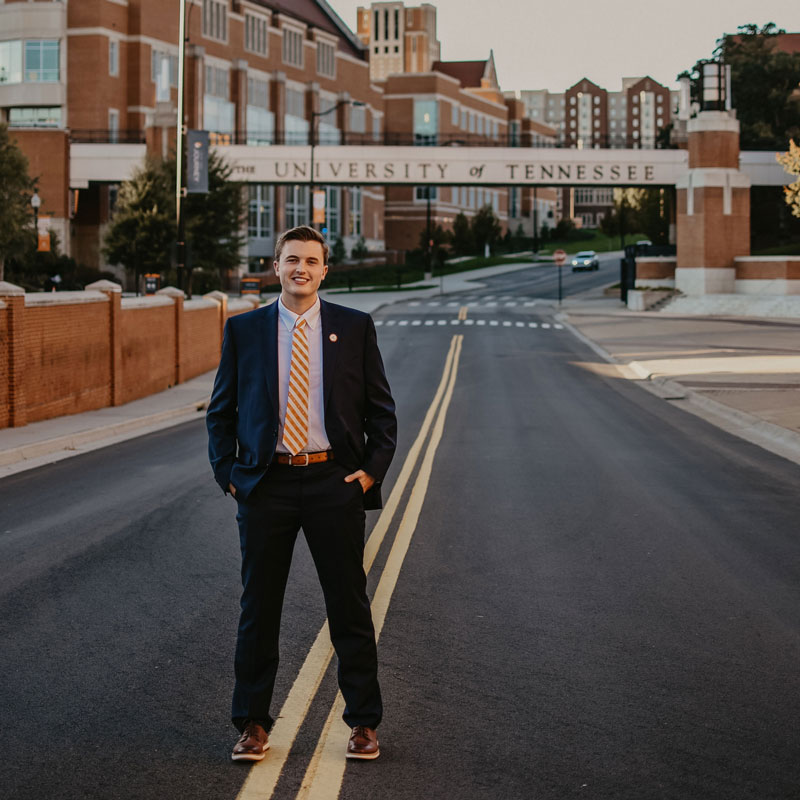 Cohl Morgan, dressed in a suit with an orange tie, stands on campus with Ped Walkway behind him.