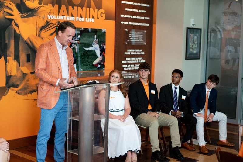peyton-manning-scholars-3-800x533 Peyton Manning stands at a lectern, addressing attendees of the Peyton Manning Scholars reception as the four 2025 scholars look on.
