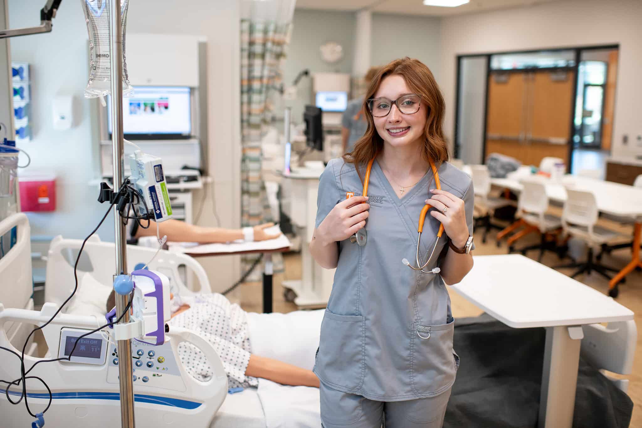 2026-Nursing-Hannah-Moody-8718 Nursing in front of hospital bed smiling in blue scrubs