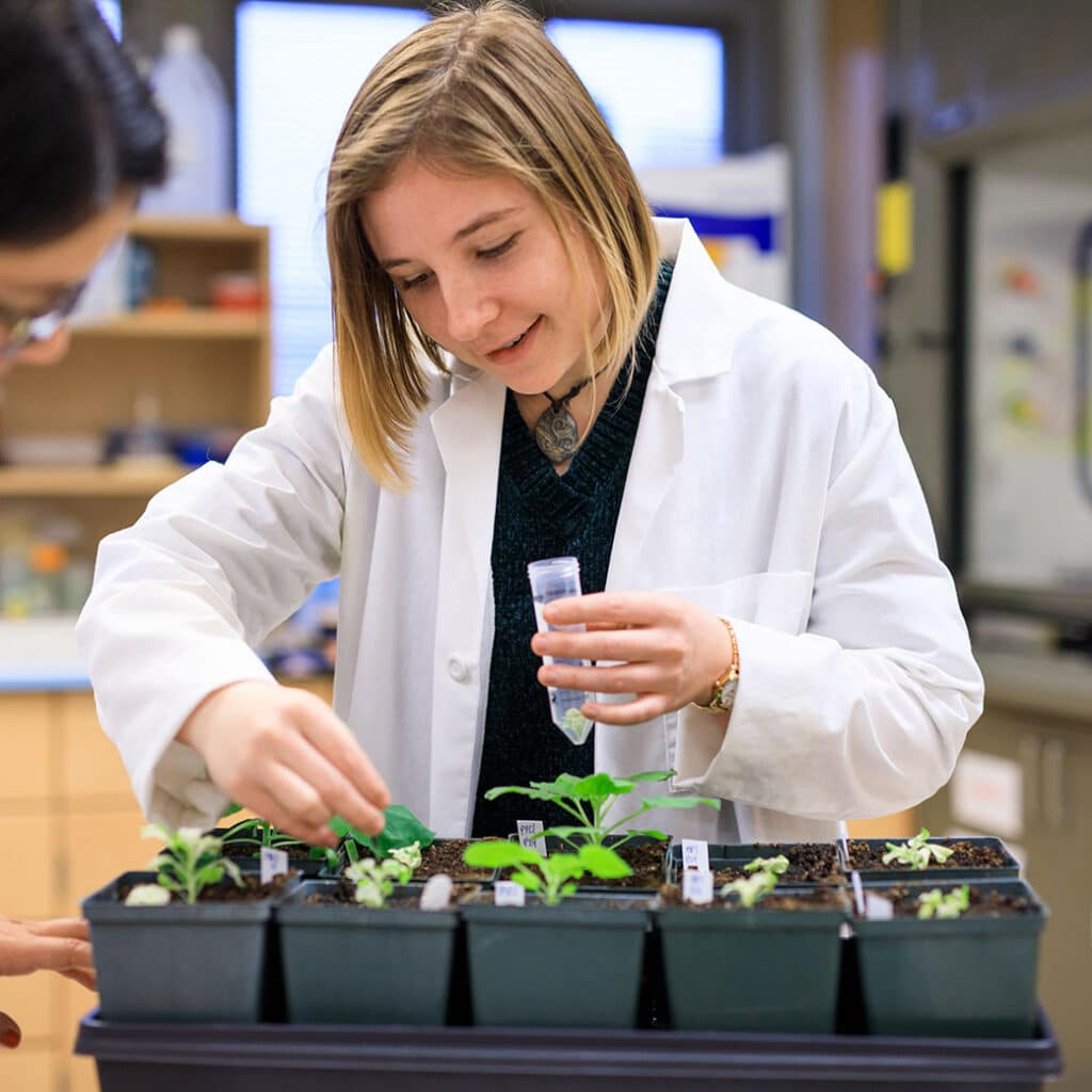 Student in white coat with beaker working with plants